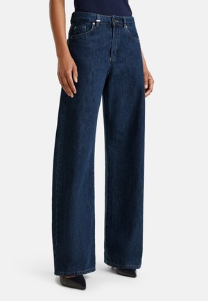 Un jean en denim large bleu foncé porté par une personne debout, associé à des chaussures noires à bout pointu et un haut sombre.