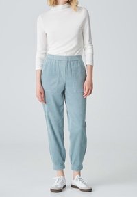 Femme portant un haut à manches longues blanc, un pantalon en maille côtelée bleu clair avec une taille élastique et des poches avant, et des baskets blanches, se tenant devant un fond uni.