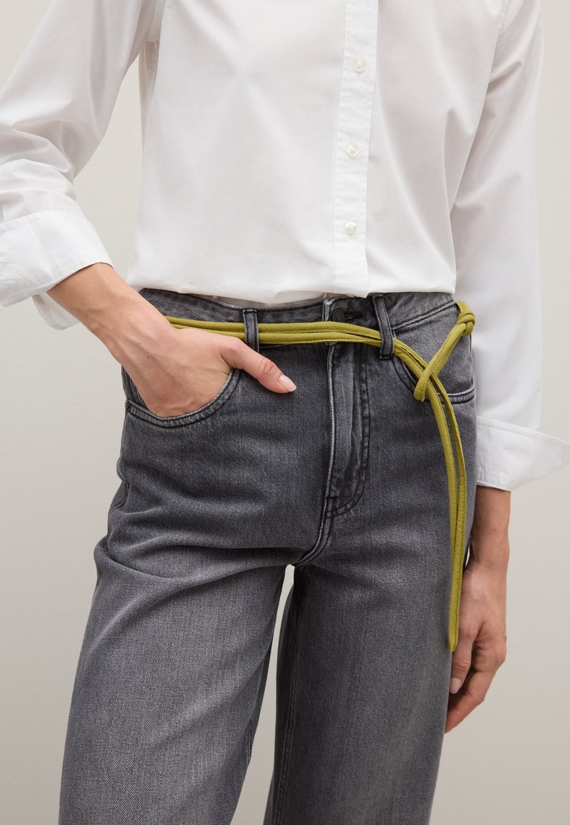 Grijze spijkerbroek wordt gedragen met een wit overhemd met knopen, voorzien van een fel limoen groene riem die om de taille is gestrikt, wat een contrasterend accent toevoegt.