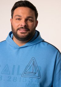 Ljusblå hoodie med dragsnöre, prydd med en segelbåtsgrafik i svart. Mjuk tyg med en slät yta och sidodetaljer i randigt.