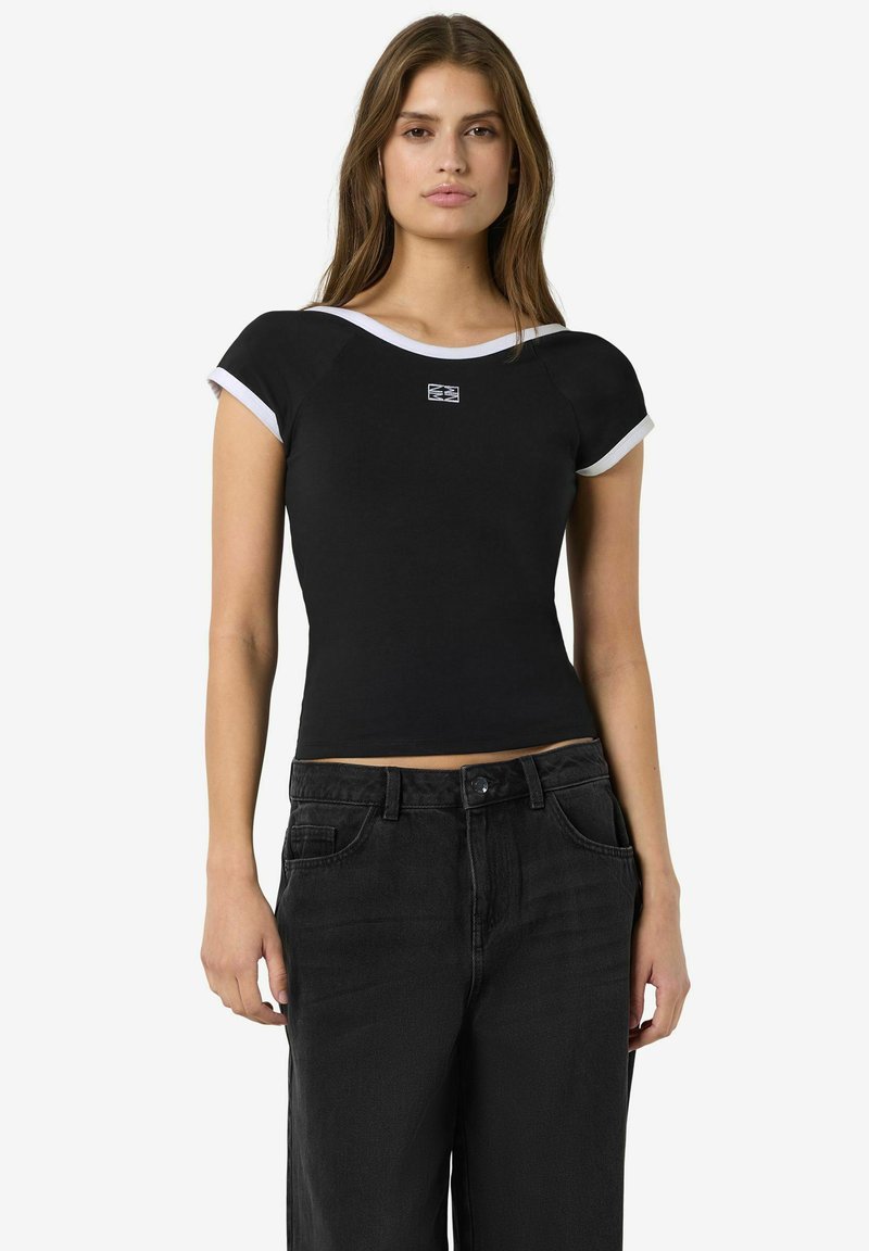 Femme portant un haut noir moulant à manches courtes avec un liseré blanc et un jean noir ample taille haute, debout devant un fond uni.