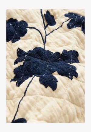 Tkanina pikowana w odcieniu beżowym z misternym granatowym haftem kwiatowym. Posiada teksturowane warstwy i widoczne detale szwów w powtarzającym się wzorze.