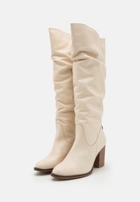 Botas de rodilla color crema con un diseño arrugado, hechas de cuero suave. Presentan una puntera cuadrada y un tacón de madera apilada.