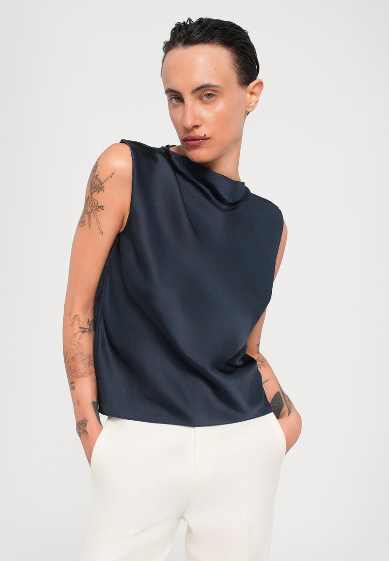 Marine satin ærmeløs top med høj hals og draperet design, parret med hvide skræddersyede bukser. Har en glat tekstur og minimalistisk form.