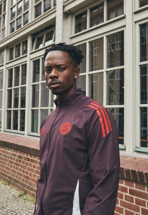 Veste de sport bordeaux avec des accents rouges, fermeture éclair et logo. Présente trois bandes rouges sur chaque épaule et des panneaux latéraux blancs.