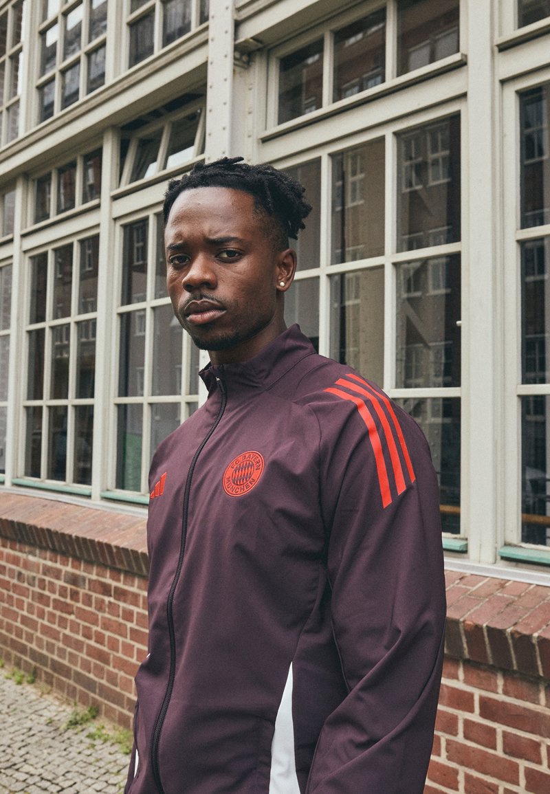 Giacca sportiva bordeaux con dettagli rossi, chiusura con zip e logo. Presenta tre strisce rosse su ciascun spalla e pannelli laterali bianchi.