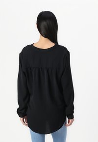 Camisa negra de manga larga con corte holgado, con un escote redondo, detalle recogido en la parte trasera y un dobladillo ligeramente curvado.