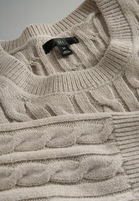 Bežani pleteni pulover s teksturiranim kabel dizajnom, rebrastim ovratnikom i oznakom s veličinom "M." Mekana tekstura s višeslojnim izgledom.