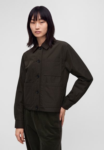 Femme aux cheveux noirs et raides porte une veste boutonnée sombre avec trois poches avant et un pantalon en velours côtelé foncé.