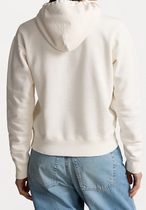 Osoba ubrana w gładką, kremowo-białą bluzę z kapturem i jasnoniebieskie dżinsy, pokazana od tyłu z rękami opuszczonymi wzdłuż ciała.