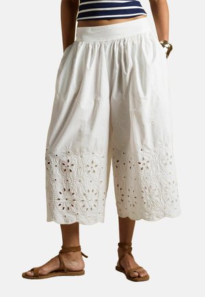 White cropped wide-leg pants with floral eyelet embroidery and scalloped hem, paired with brown ankle-strap sandals and a striped top.