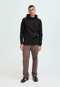 Sudadera negra con bolsillo tipo canguro, capucha con cordón, mangas largas y puños acanalados. Combinada con pantalones marrones y zapatillas negras.