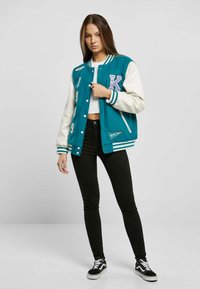 Veste universitaire bleu-vert avec des manches crème, ornée de lettres et de chiffres brodés. Associée à un jean skinny noir et des baskets noir et blanc.