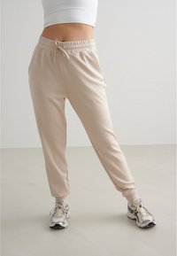 Beige Sweatpants mit elastischem Taillenbund, Kordelzug und Seitentaschen, kombiniert mit grau-weißen Sneakers. Glatte Stoffoberfläche.