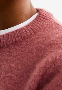 Rosa ulltröja med ribbad halsringning, som har en mjuk textur och en något lös passform. Vit lager synlig under.