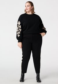 Zwarte sweatshirt met geborduurd bloemenpatroon op de mouwen, gecombineerd met zwarte broek met zijstrepen en zwarte enkellaarzen.