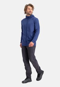 Navy zip-up jacket with hood, made of smooth fabric. Paired with dark pants and black footwear featuring a textured sole.