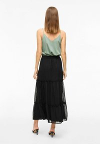 Camisole vert en v associé à une jupe noire à volants. La jupe présente une texture en tulle à pois et une taille élastique. Sandales à talons visibles.