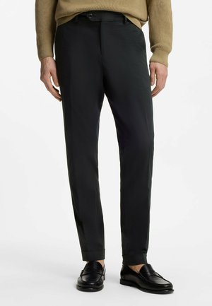 Uomo che indossa pantaloni neri slim fit con orli risvoltati e mocassini in pelle nera, abbinati a un maglione marrone, in piedi su sfondo bianco.