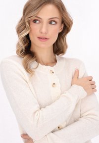 Cardigan crem din țesătură tricotață, cu un decolteu în valuri, două butoane mari și manșete cu dungi. Textură moale și o senzație ușoară.