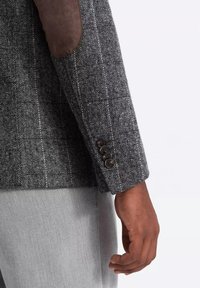 Blazer grigio a spina di pesce con toppe in suede marrone sui gomiti. Presenta un polsino a quattro bottoni. Il tessuto ha una finitura texturizzata e un design strutturato.