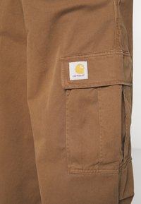 Nahaufnahme von einer braunen Carhartt-Hosentasche mit sichtbar eingewebtem Markenlogo auf dem Stoffdeckel.