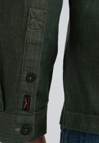 Chemise en chevrons vert foncé avec des boutons aux poignets et une étiquette de marque discrète sur la manche. Le tissu a un aspect texturé et une coupe structurée.