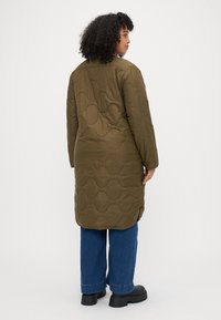 Μακρύ, ελαιόλαδο πράσινο καπιτονέ παλτό με κυματιστό σχέδιο, διαθέτοντας μακριά μανίκια και πλαινές σχισμές, συνδυασμένο με μπλε φαρδιά τζιν και μαύρες χοντρές μπότες.