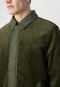 Olive green corduroy jacket with a textured surface, collared neckline, and snap button closure. Features embroidered branding along the side.