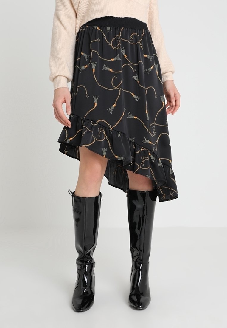 Jupe noire asymétrique avec motif chaîne et pompons, associée à un pull beige et des bottes noires brillantes montant jusqu'aux genoux.