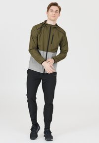Oliivinvihreä ja harmaa kevyt takki, jossa on huppu ja vetoketjullinen tasku, yhdistettyinä mustiin slim-fit-housuihin ja mustiin lenkkareihin.