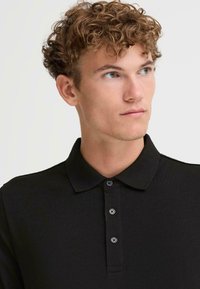 Čierne polo tričko s rebrovanou textúrou, s gombíkovou légou, klasickým golierom a krátkymi rukávmi. Modelka má kučeravé vlasy.