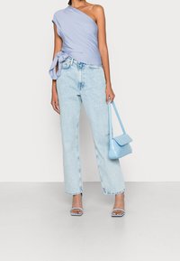 Vrouw in een lichtblauw één-schoudertop, lichtgekleurde high-waisted jeans, blauwe haksandalen en met een kleine blauwe handtas.