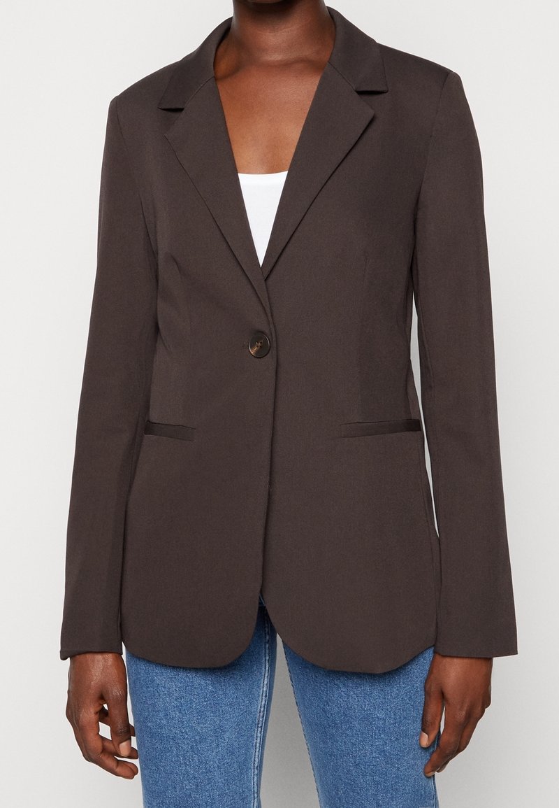 Blazer marron foncé à un seul bouton porté sur un top blanc et un jean bleu, visible de la poitrine jusqu’aux hanches sur une personne à la peau foncée.