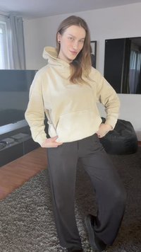 Cream-colored fleece hoodie with a front pocket, paired with loose-fitting black pants. Soft texture and relaxed design, set in a modern indoor space.