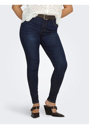 Kvinde iført mørkeblå skinny jeans, en hvid eyelet bluse, brun bælte og sorte kilehæle med spids tå og ankelremme.