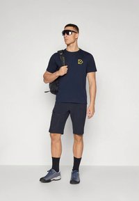 Μπλε ναυτικό T-shirt με κίτρινο λογότυπο, μαύρο σορτς και παπούτσια πεζοπορίας. Άτομο που φορεί μαύρο σακίδιο πλάτης, γυαλιά ηλίου και σκούρες κάλτσες.