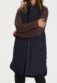 Gilet senza maniche trapuntato navy con chiusura a bottoni, orlo arrotondato e due tasche laterali. Indossato sopra un maglione lavorato a maglia spesso di colore bordeaux.