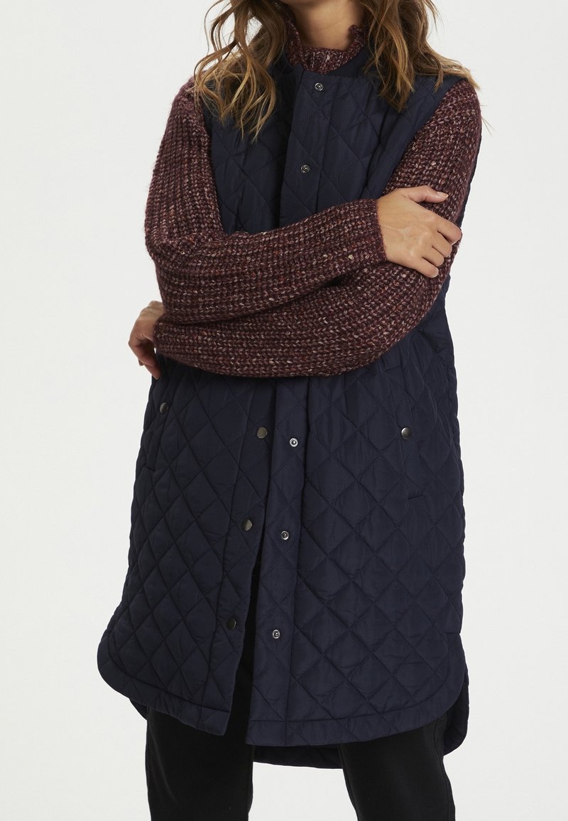 Gilet senza maniche trapuntato navy con chiusura a bottoni, orlo arrotondato e due tasche laterali. Indossato sopra un maglione lavorato a maglia spesso di colore bordeaux.