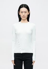 Biely top s dlhými rukávmi vyrobený z elastickej látky s okrúhlym výstrihom a asymetrickým lemom so striebornými patentkami na boku.