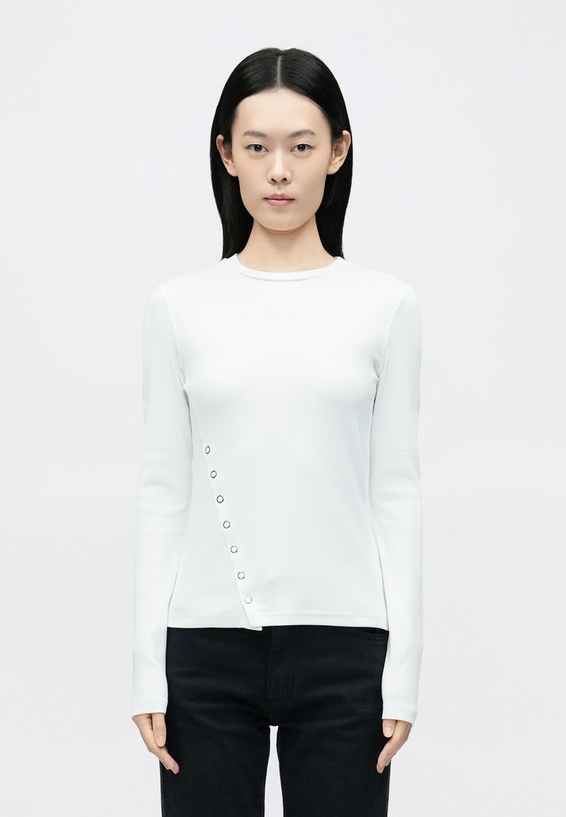 Biely top s dlhými rukávmi vyrobený z elastickej látky s okrúhlym výstrihom a asymetrickým lemom so striebornými patentkami na boku.