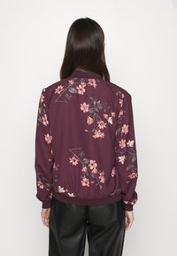 Burgundy květinová bomber bunda s růžovými květy, elastickými manžetami a lemováním. Hladká tkanina a volný střih. Zobrazený zadní pohled.