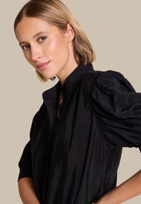 Svart skjorte med høy krage og puffede ermer, laget av et glatt stoff med en subtil glans. Bakgrunnen er nøytral beige.