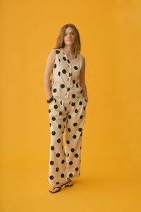 Femme debout sur un fond jaune portant un haut beige sans manches boutonné et un pantalon large à gros pois noirs, mains dans les poches.