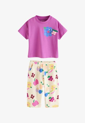 Dwuczęściowy strój dziecięcy z fioletową bluzką z krótkim rękawem zdobioną uśmiechniętym obrazkiem jagody oraz kremowymi spodniami z kolorowymi wzorami owoców.