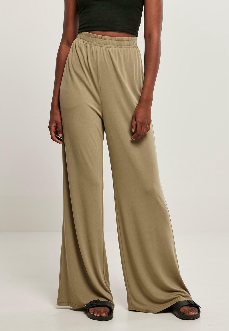 Personne portant un pantalon beige ample taille haute, un crop top noir et des sandales noires, debout devant un fond clair uni.