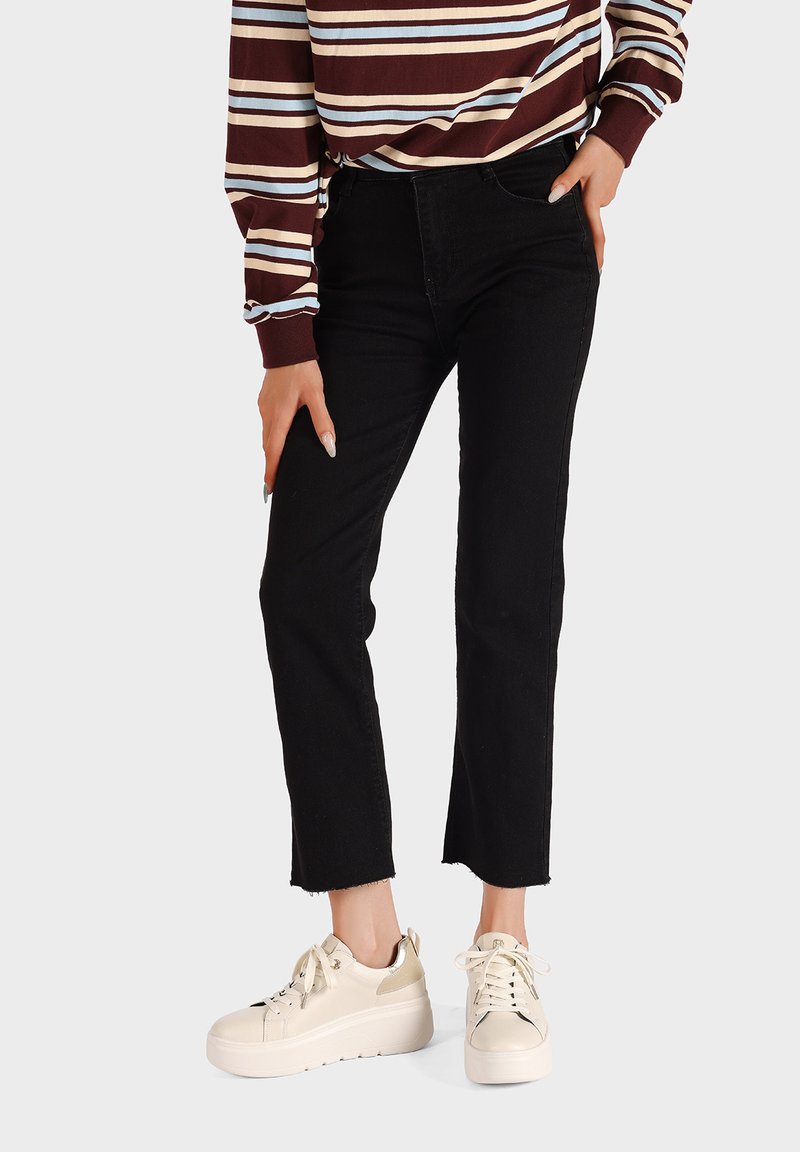 Jeans in denim neri con orlo sfrangiato, indossati con sneakers beige con piattaforma. L'outfit presenta un top a maniche lunghe a righe bordeaux e crema.