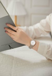 Reloj de pulsera plateado y oro rosa con esfera redonda y dial blanco. La correa tiene un diseño de metal eslabonado. Una mano sosteniendo un libro gris reposa sobre una mesa texturizada.
