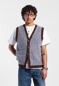 Gilet en tricot bleu et marron avec une texture côtelée, design en V, cinq boutons, porté sur un T-shirt blanc, associé à un jean en denim.