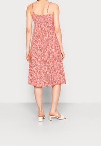 Rotes Midi-Kleid mit weißen Punkten, taillierter Oberkörper und gerafftem Rücken. Dünne Träger führen zu einem fließenden Rock. Kombiniert mit crèmefarbenen Blockabsatzsandalen.
